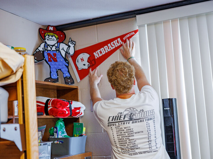 a student hanging a Nebraska Huskers pennant in his residence hall room