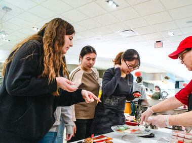 Students gather around a Lunar New Year sample plate as a Dining Services team member explains the food being offered.