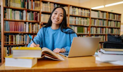 Student studying in a library with books and a laptop on the table in front of them